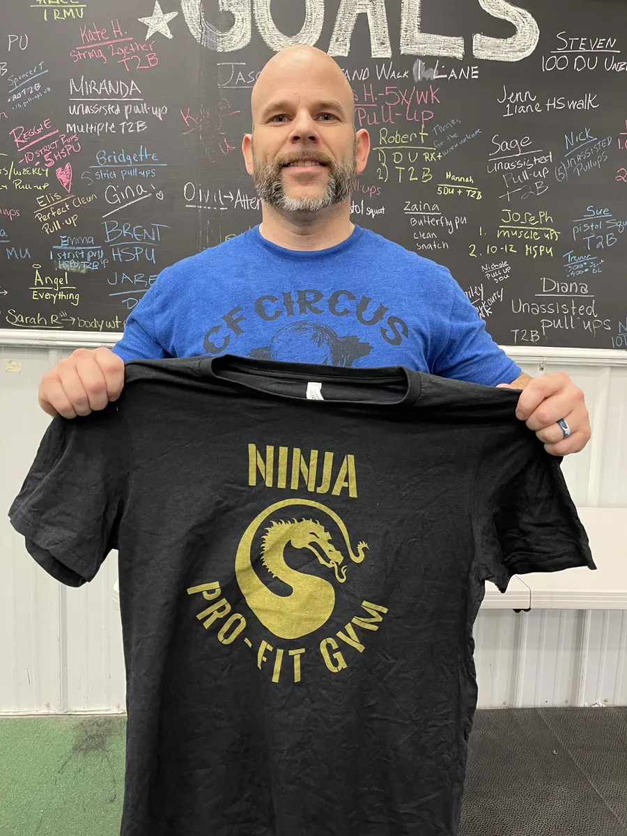 A member holds up his earned Ninja shirt in front of the member goals board